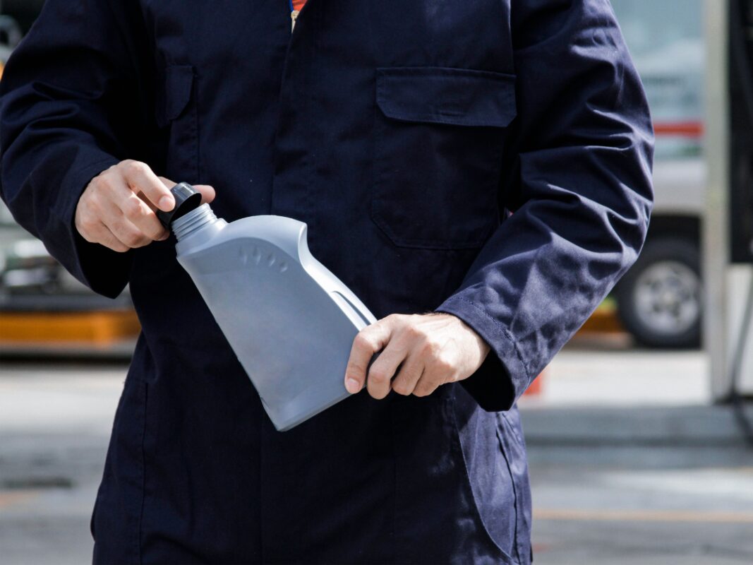 Close up photo of a person in a blue mechanic jumpsuit holding a label-free motor oil bottle.
